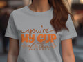 A person wears a 100% cotton graphic T-shirt from Garment Graphics, with orange text saying Youre My Cup of Tea and small tea cups. Their long, wavy hair partially conceals their face against a blurred background.