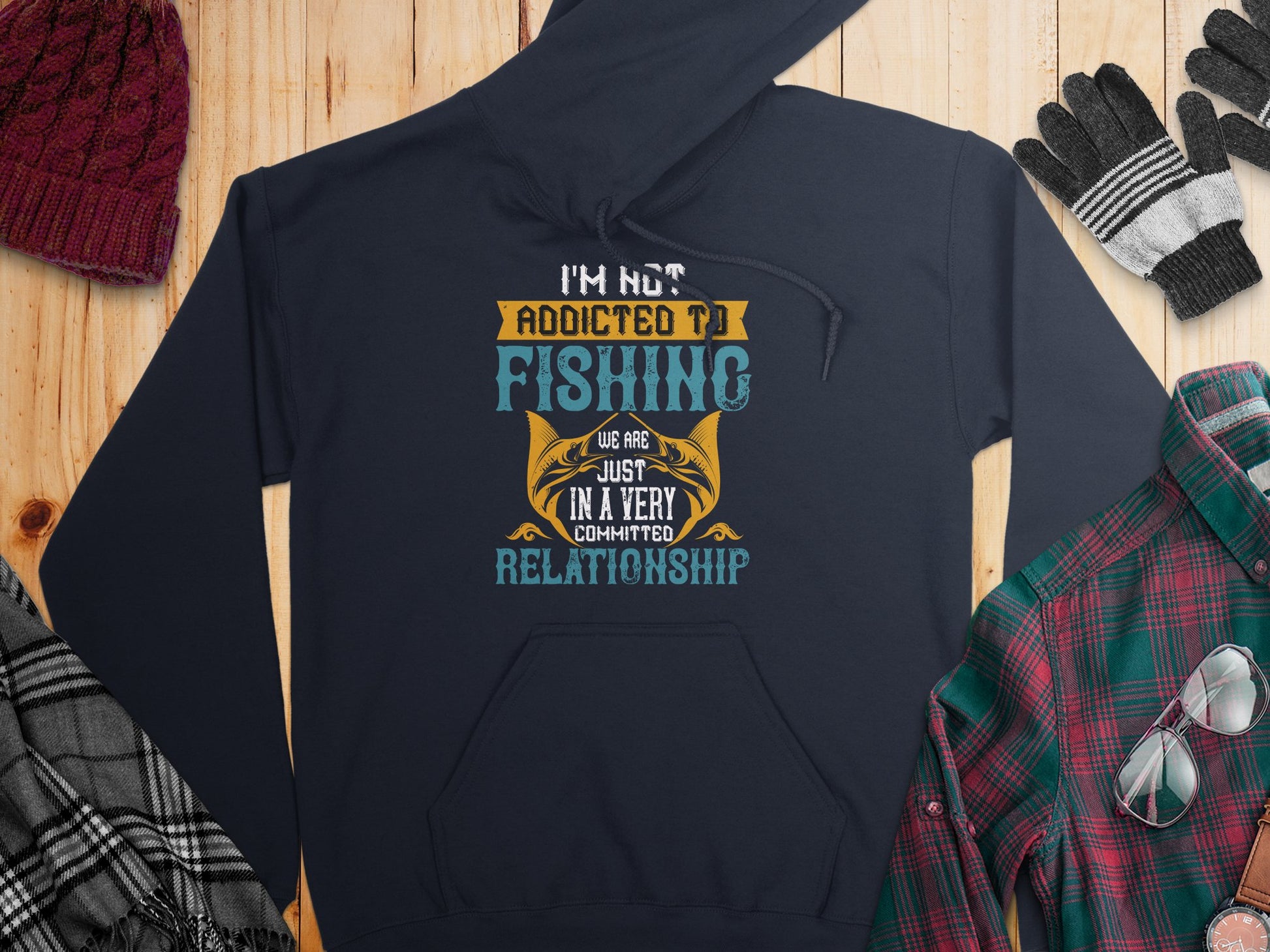 A navy fishing hoodie by Garment Graphics features the phrase Im not addicted to fishing, we are just in a very committed relationship. It complements winter staples like a maroon knit hat, plaid scarf, brown boots, plaid shirt, gloves, and sunglasses on a wooden surface.