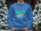 Someone wearing a Garment Graphics blue sweatshirt, which features Take It Easy with arrows and musical notes, stands comfortably among snow-covered trees. This vibrant design is available in unisex sizing.