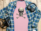 A flat lay image presents a striking look with Garment Graphics pink graphic t-shirt featuring a skull and crossbones. Its accompanied by a blue plaid shirt, headphones, a camera, and a takeaway coffee cup on a wooden surface.