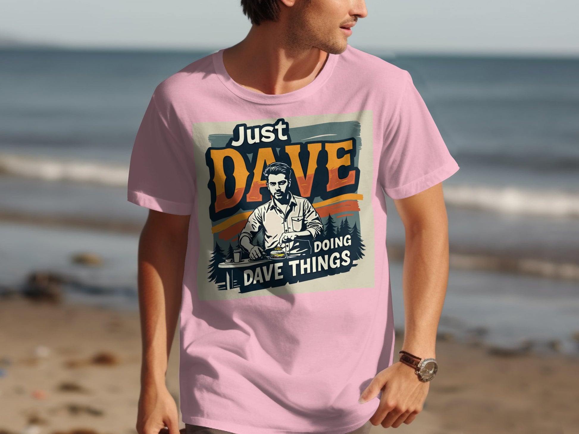 A man stands on a beach wearing a Garment Graphics classic fit pink T-shirt with the Just Dave Doing Dave Things graphic design, set against the sea and sand backdrop.