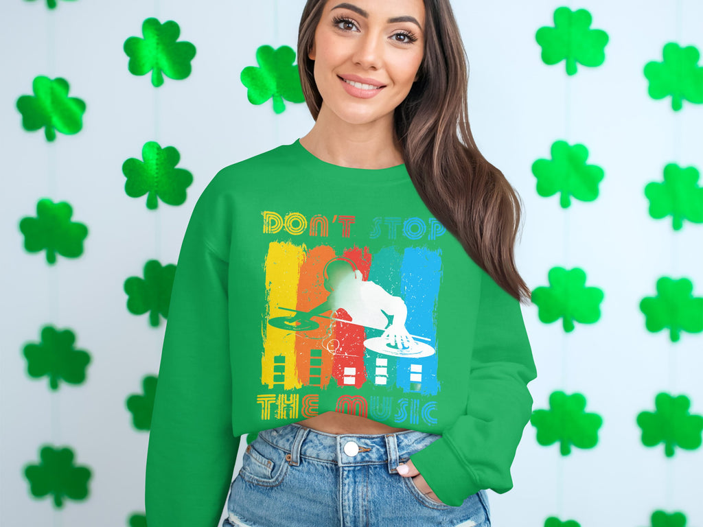 A music lover in a Garment Graphics green sweater with a DJ design and Dont Stop the Music stands before a backdrop of green shamrocks, smiling warmly with long hair cascading, hands in denim pant pockets.