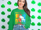 A music lover in a Garment Graphics green sweater with a DJ design and Dont Stop the Music stands before a backdrop of green shamrocks, smiling warmly with long hair cascading, hands in denim pant pockets.