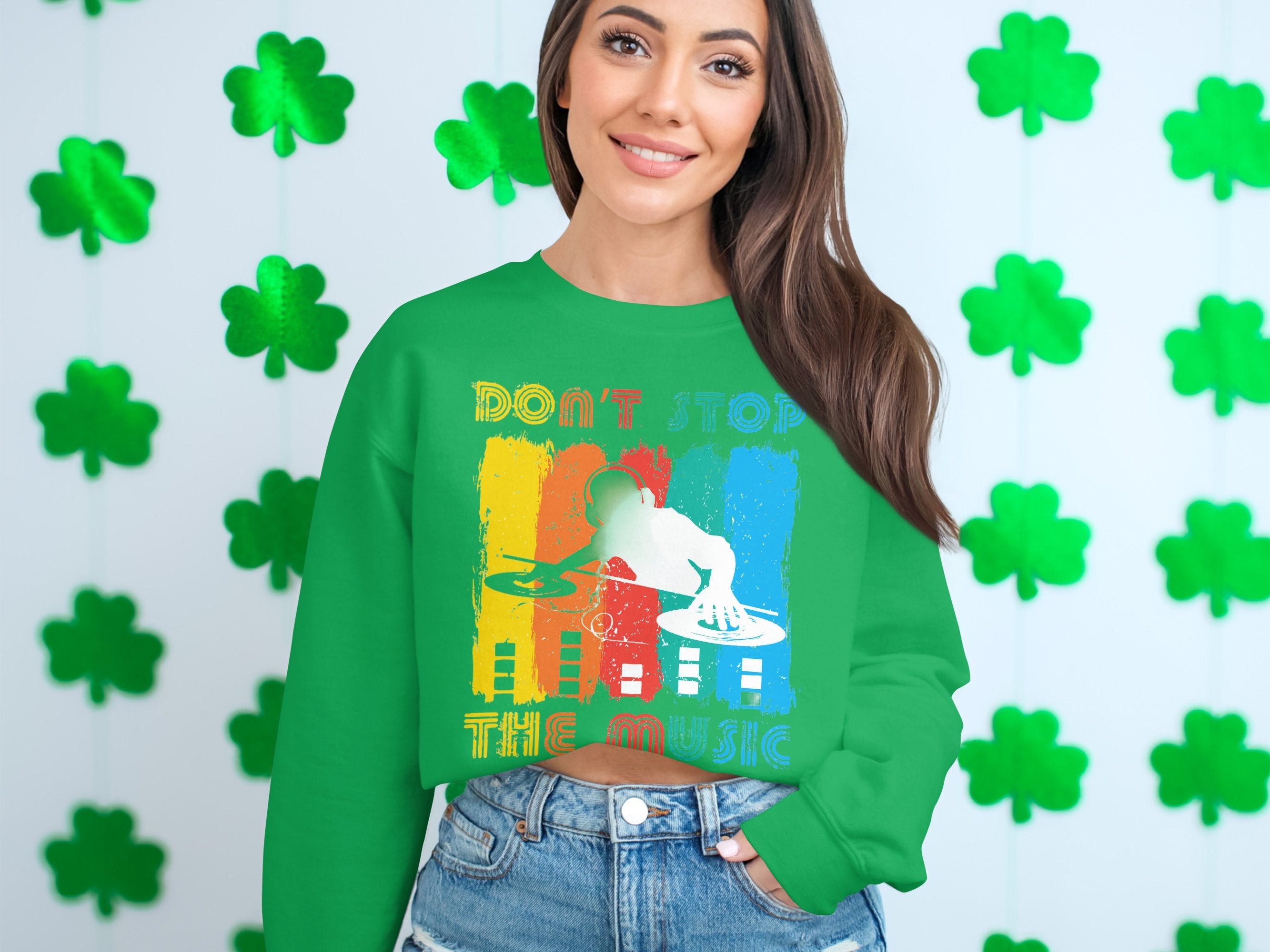 A music lover in a Garment Graphics green sweater with a DJ design and Dont Stop the Music stands before a backdrop of green shamrocks, smiling warmly with long hair cascading, hands in denim pant pockets.