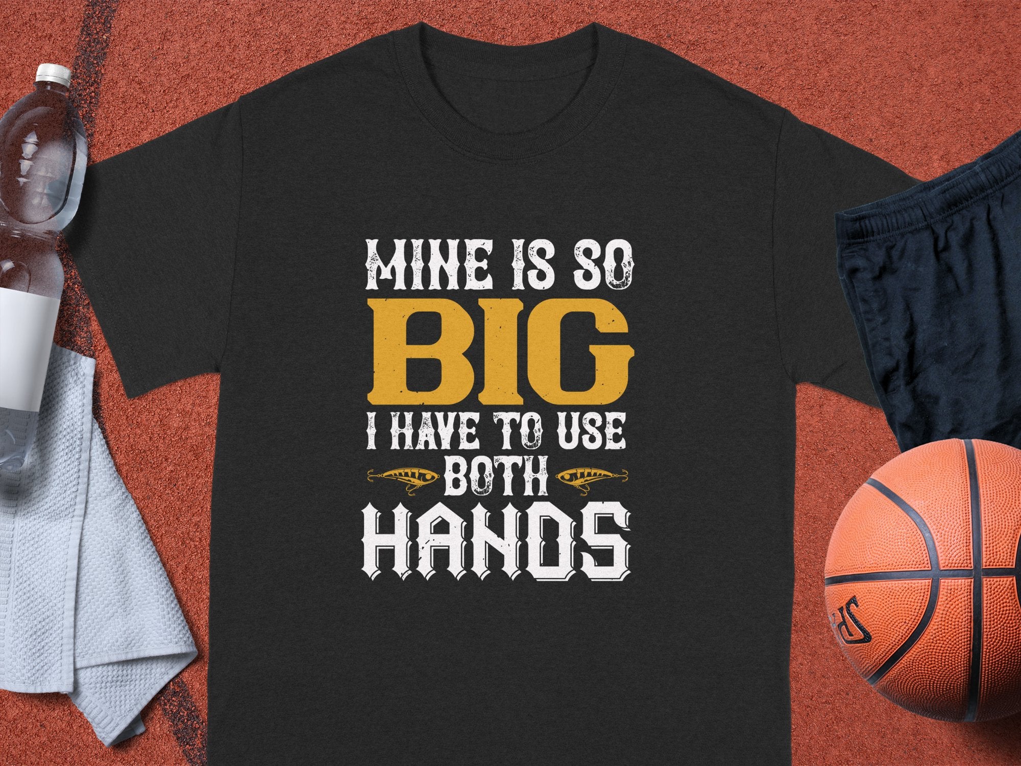 A black graphic tee from Garment Graphics lies on a sports court, featuring the humorous design: Mine is so big I have to use both hands with two small football graphics. Nearby are a water bottle, white towel, and basketball, emphasizing the classic fit of this playful shirt.