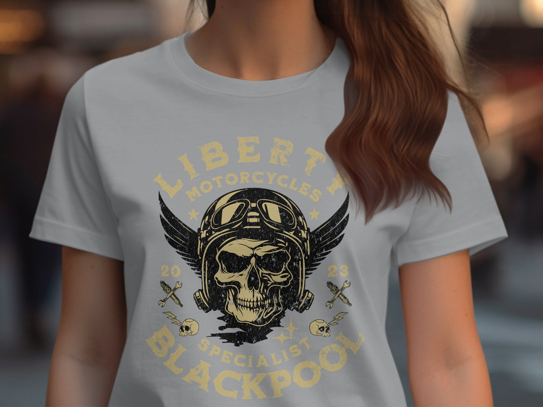 A Blackpool-themed white t-shirt from Garment Graphics features a skull with wings, pilot helmet, goggles, and Liberty Motorcycles Specialist Blackpool 2023. Its artistically displayed on a wooden surface with festive decor and a cozy sweater.