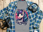 The Garment Graphics gray t-shirt, showcasing a vibrant ponyunicorn design with I love my pony, is displayed on a wooden surface. Nearby are a blue and green plaid shirt, black headphones, and a sleek black camera.