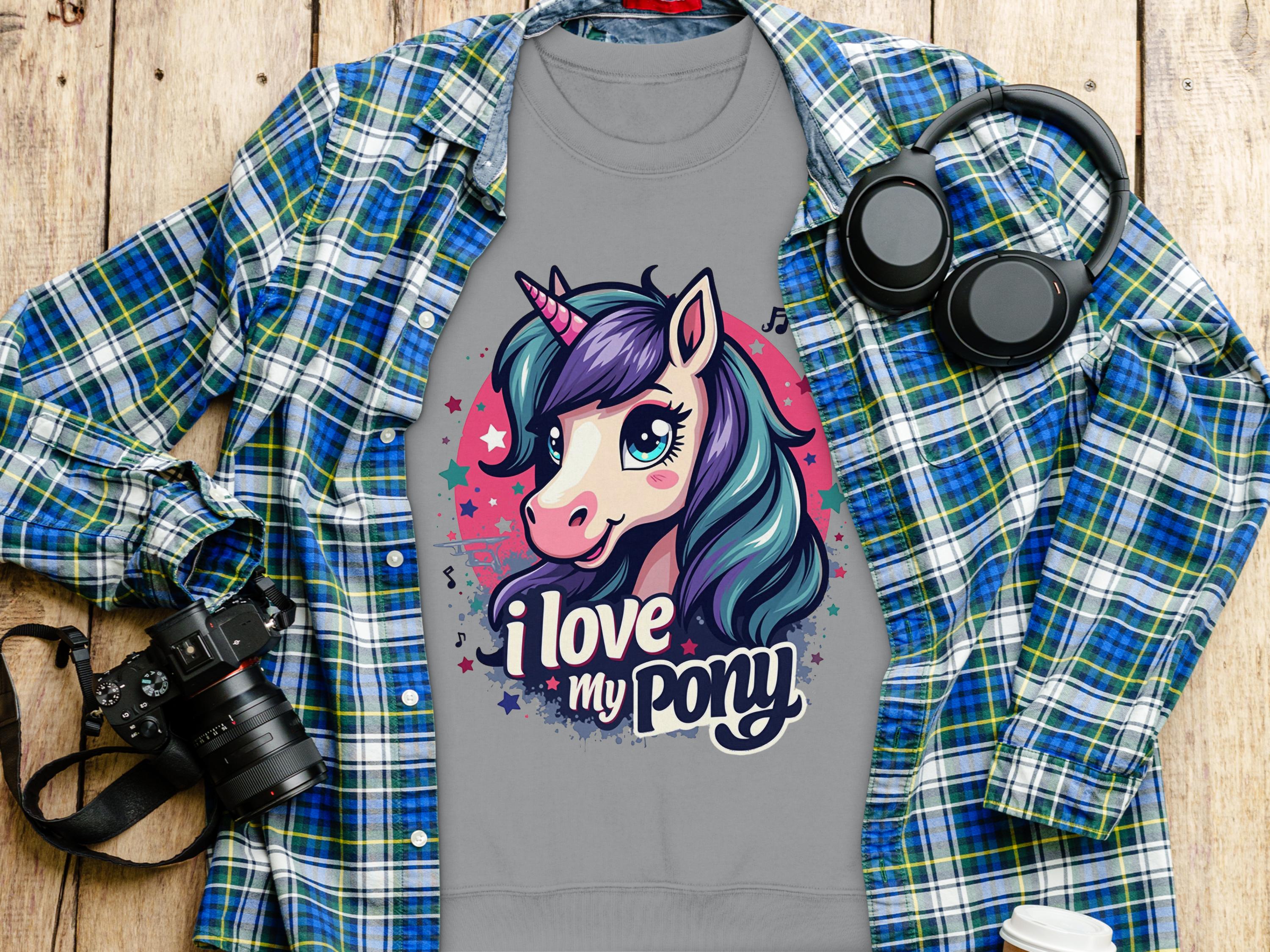 The Garment Graphics gray t-shirt, showcasing a vibrant ponyunicorn design with I love my pony, is displayed on a wooden surface. Nearby are a blue and green plaid shirt, black headphones, and a sleek black camera.