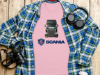 A pink Scania truck graphic T-shirt by Garment Graphics is on a wooden surface, topped with a blue and green plaid shirt. Black headphones, a camera, and a white takeaway cup nearby create the perfect scene for truck enthusiasts.