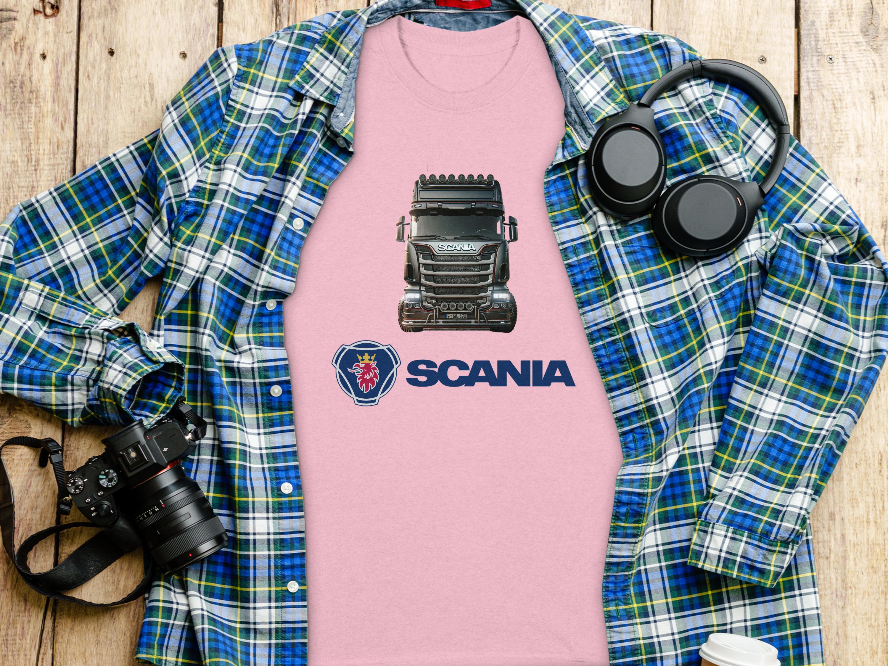A pink Scania truck graphic T-shirt by Garment Graphics is on a wooden surface, topped with a blue and green plaid shirt. Black headphones, a camera, and a white takeaway cup nearby create the perfect scene for truck enthusiasts.