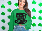 A person wearing a green St. Patricks Day-themed sweater by Garment Graphics, featuring a black hat design, exudes festive spirit in front of a wall with green shamrocks, paired with denim shorts.