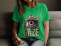 A person sits on a sofa, displaying a womens green Garment Graphics T-shirt. It features an embellished eye design surrounded by jewels, with GAZE printed below.