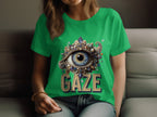 A person sits on a sofa, displaying a womens green Garment Graphics T-shirt. It features an embellished eye design surrounded by jewels, with GAZE printed below.
