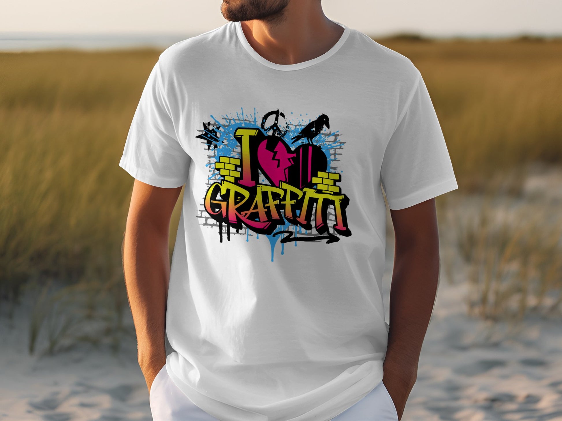 A person wears a white Garment Graphics Graffiti Love T-Shirt with vibrant I Love Graffiti text. The backdrop of sandy dunes and tall grass beneath a partly cloudy sky captures the dynamic spirit of street art.
