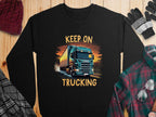 Garment Graphics sweatshirt, ideal for truck enthusiasts, features a large semi-truck illustration on a road with clouds and KEEP ON TRUCKING boldly printed above. Its styled with a hat, gloves, and plaid shirt on a wooden surface.