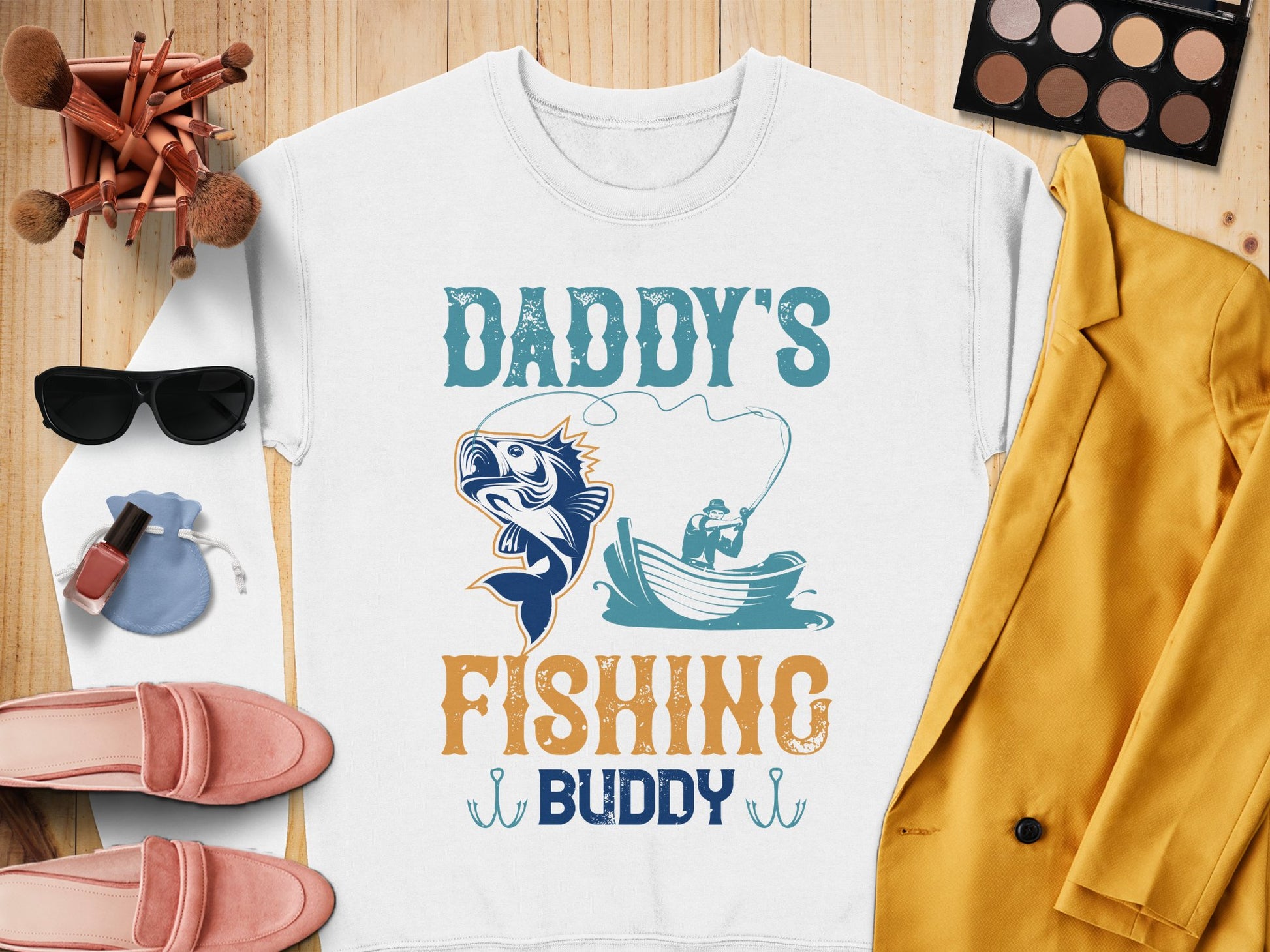 The white t-shirt by Garment Graphics features a person fishing with Daddys Fishing Buddy text. Surrounded by accessories like sunglasses and makeup, its perfect for casual wear or paired with cozy fishing sweatshirts in cooler weather. Ideal for any fishing enthusiast!.