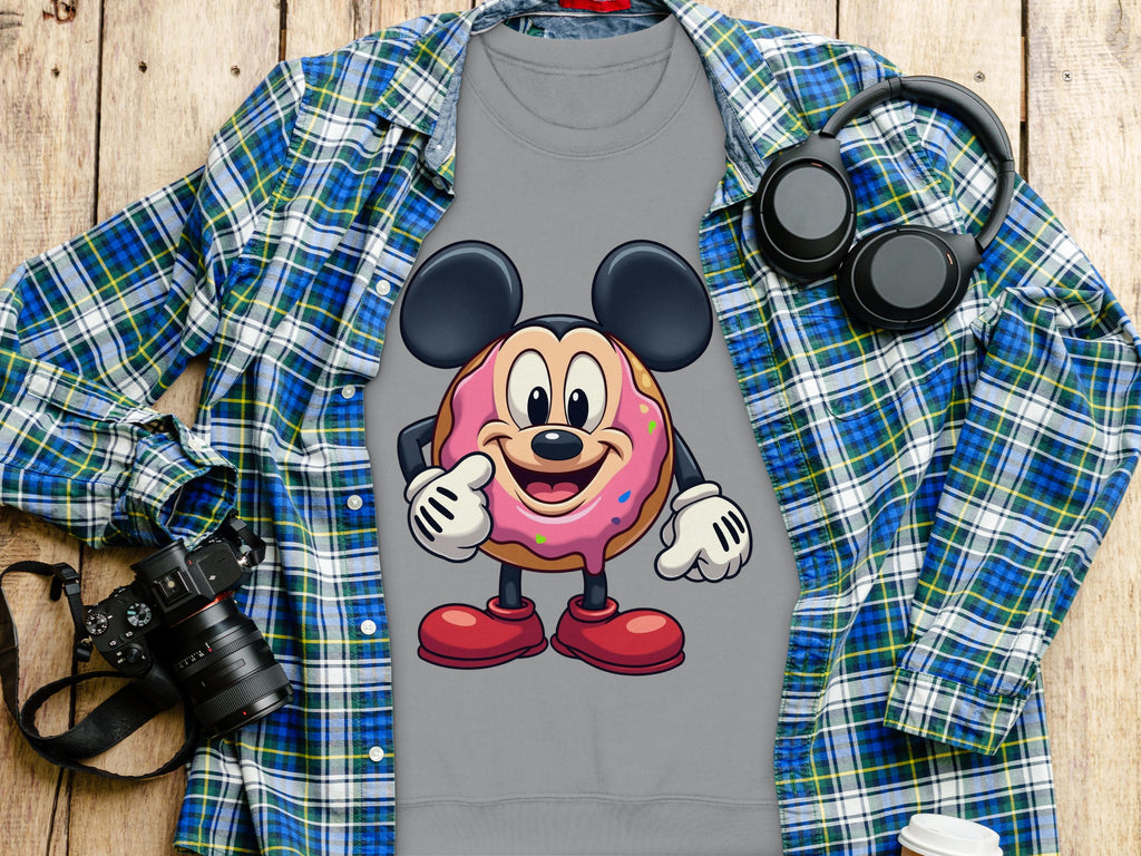 A Garment Graphics gray t-shirt showcases a cute cartoon donut with mouse ears and red shoes on pink icing. Paired with a blue-green plaid shirt, it radiates playfulness. Nearby on the wooden surface are a camera, headphones, and a disposable coffee cup.