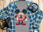A Garment Graphics gray t-shirt showcases a cute cartoon donut with mouse ears and red shoes on pink icing. Paired with a blue-green plaid shirt, it radiates playfulness. Nearby on the wooden surface are a camera, headphones, and a disposable coffee cup.