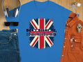 A Garment Graphics blue T-shirt, in classic fit, features a striking Union Jack design and the text Dont Blame Me, I Didnt Vote For Him, displayed next to headphones, sunglasses, a brown shirt, watch, jeans, and keys on a wooden surface.