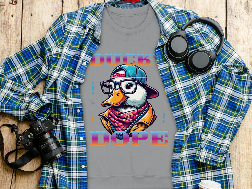 The Garment Graphics gray T-shirt features a colorful duck in a cap and glasses with DUCK DOPE text. Styled with a wooden background, blue plaid shirt, headphones, camera, and white cup—perfect for those embracing bold cool swag. Medium-heavy fabric ensures comfort and durability.