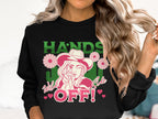 A person with long wavy hair wears a loose-fit black sweatshirt from the Garment Graphics collection, featuring unique designs of a person in a hat surrounded by flowers and hearts, with the bold phrases Hands Off and Wild Side Off!.
