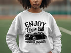 A young person sports a white Garment Graphics sweatshirt with a Classic Car graphic, stating ENJOY Classic CAR and Vintage Ride Community Aircooled Est. 1980. Amidst an outdoor blur, this stylish piece perfectly evokes the timeless charm of vintage rides.
