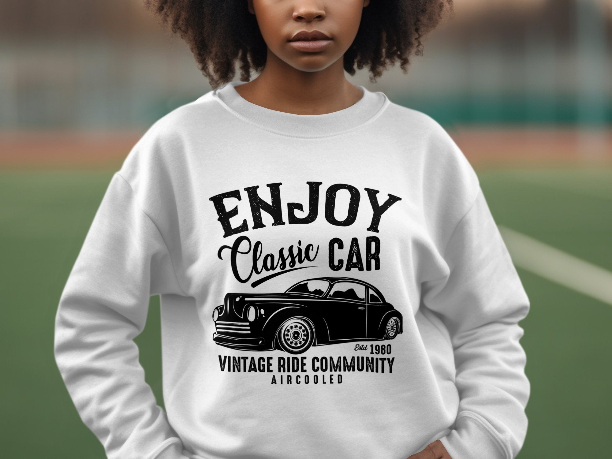 A young person sports a white Garment Graphics sweatshirt with a Classic Car graphic, stating ENJOY Classic CAR and Vintage Ride Community Aircooled Est. 1980. Amidst an outdoor blur, this stylish piece perfectly evokes the timeless charm of vintage rides.