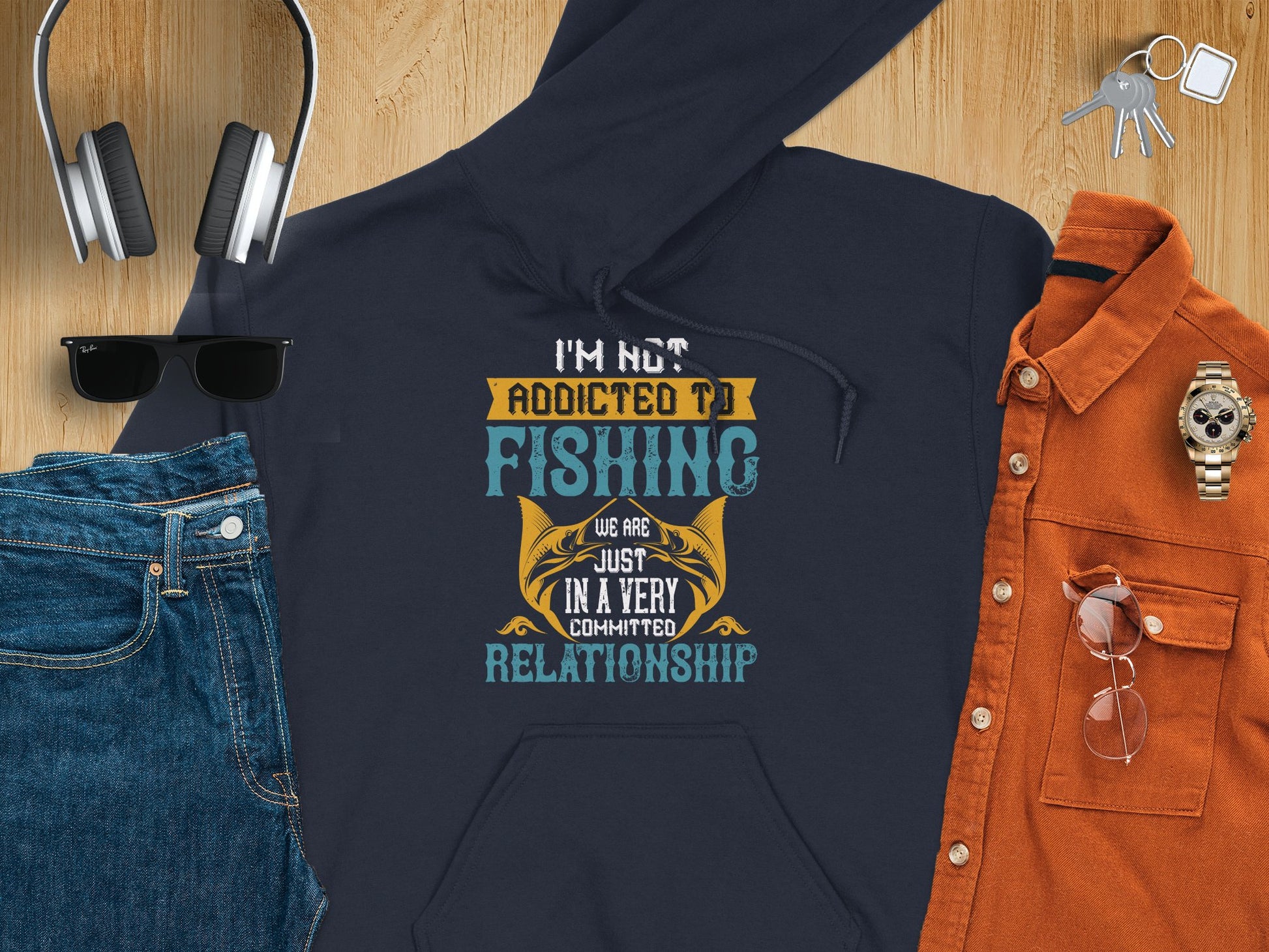 Flat lay of a classic fit navy fishing hoodie by Garment Graphics with the text, Im not addicted to fishing, we are just in a very committed relationship, surrounded by headphones, keys, sunglasses, brown button-up shirt, gold watch, glasses and blue jeans on a wooden surface.