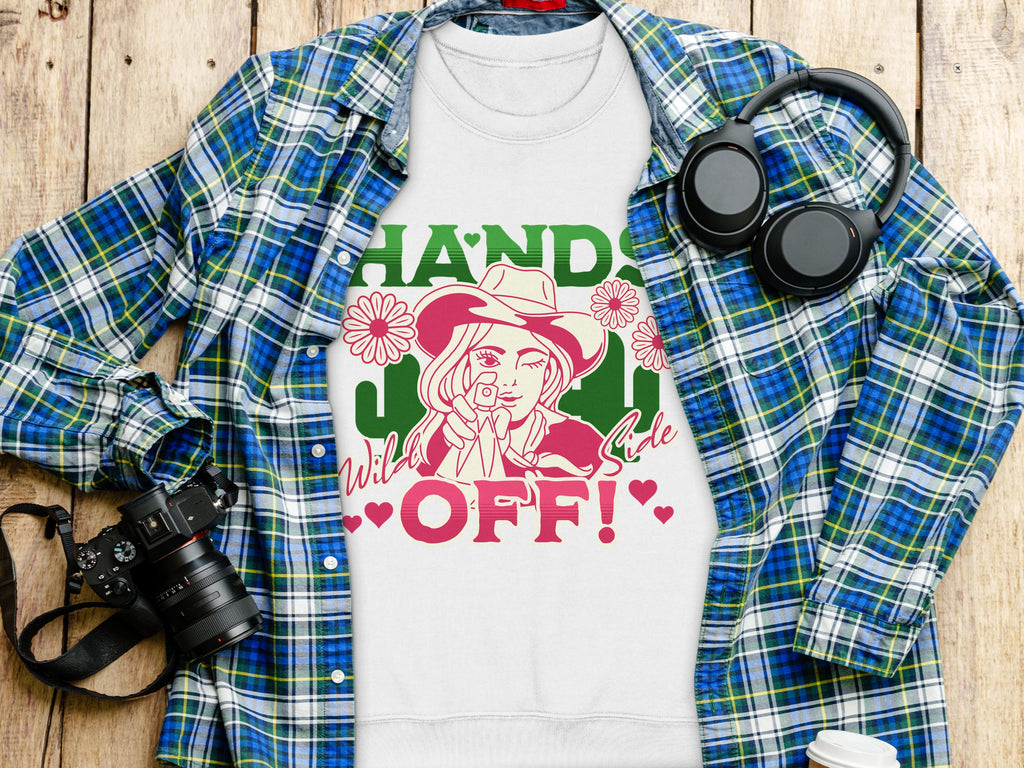 A Garment Graphics white t-shirt featuring bold imagery of a person in a hat, flowers, and Hands Off! text is showcased on wood. Nearby are a blue plaid shirt, headphones, and a camera—ideal for adventurous spirits.