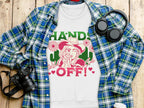 A Garment Graphics white t-shirt featuring bold imagery of a person in a hat, flowers, and Hands Off! text is showcased on wood. Nearby are a blue plaid shirt, headphones, and a camera—ideal for adventurous spirits.