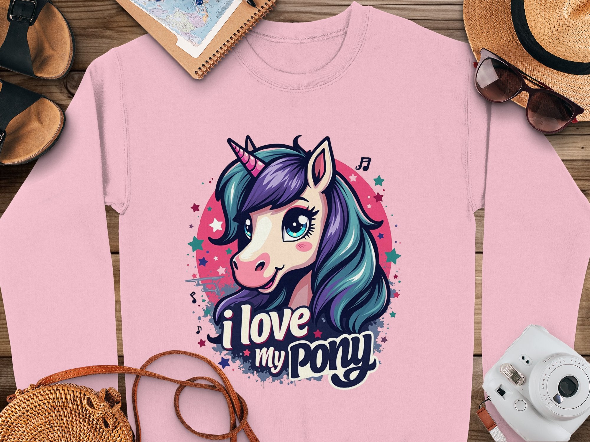 A Garment Graphics pink graphic sweatshirt with a ponyunicorn head and I love my pony text lies on wood. Paired are a notebook, sunglasses, straw hat, purse, and black sandals. The fabric features vibrant music notes and stars.