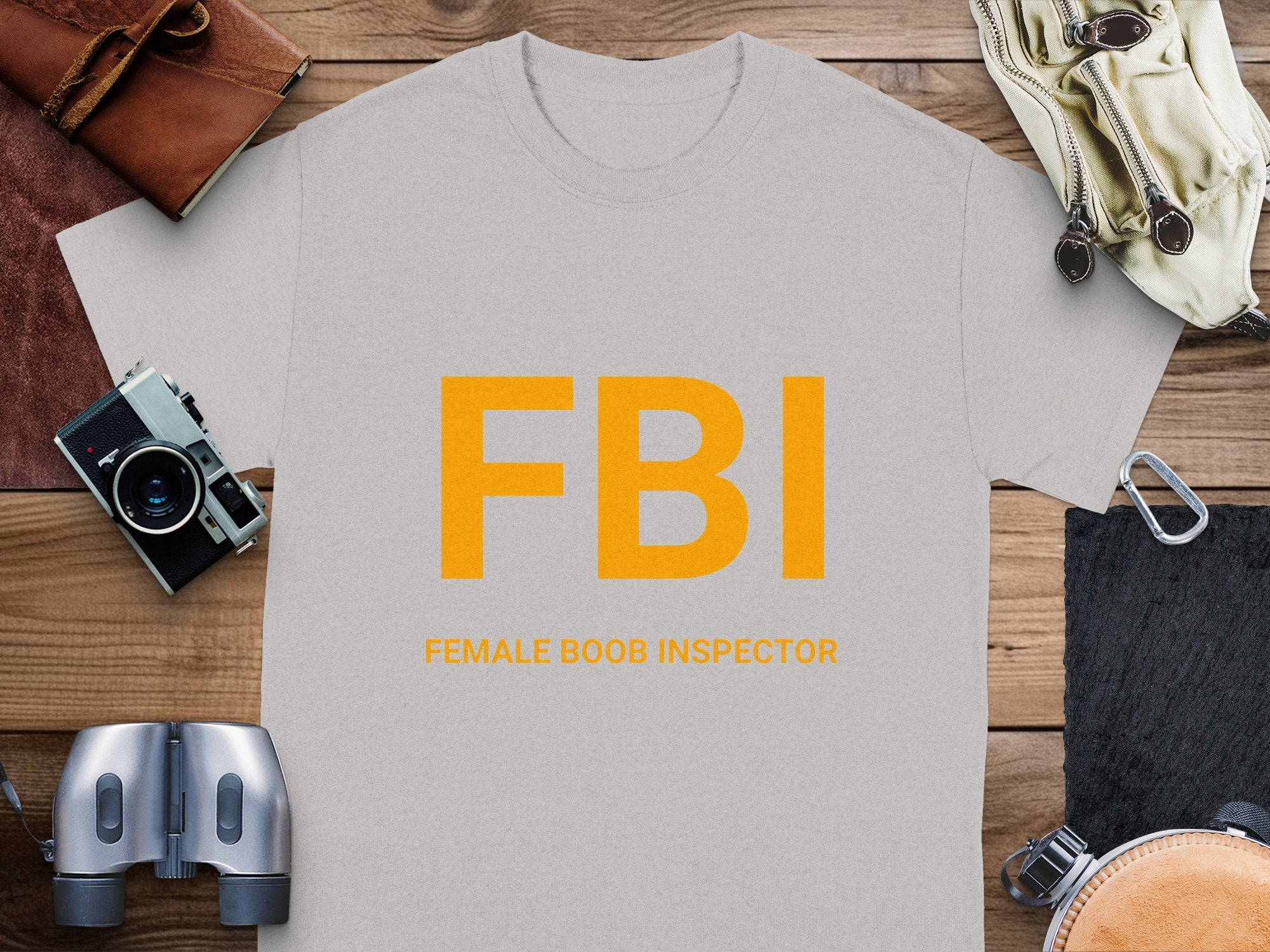 A humorous Garment Graphics T-shirt features the playful FBI Female Boob Inspector design in bold yellow letters on a wooden surface. It is complemented by a camera, leather pouch, binoculars, and gloves, creating an adventurous vibe.