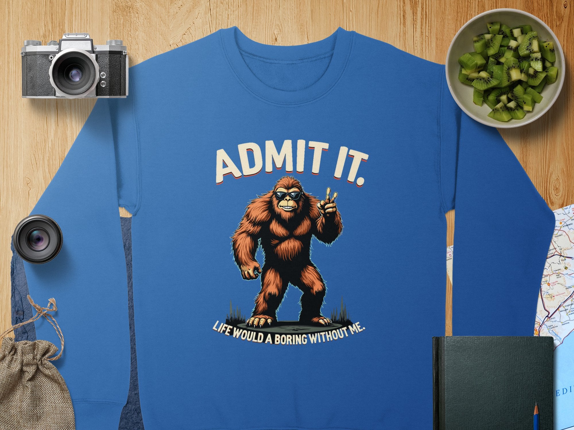 A Garment Graphics blue sweatshirt with a whimsical Bigfoot illustration and the text Admit it. Life would be boring without me. in unisex sizing, surrounded by a camera, lens, diced kiwi, map, notebook, and drawstring bag—ideal for casual wear lovers.