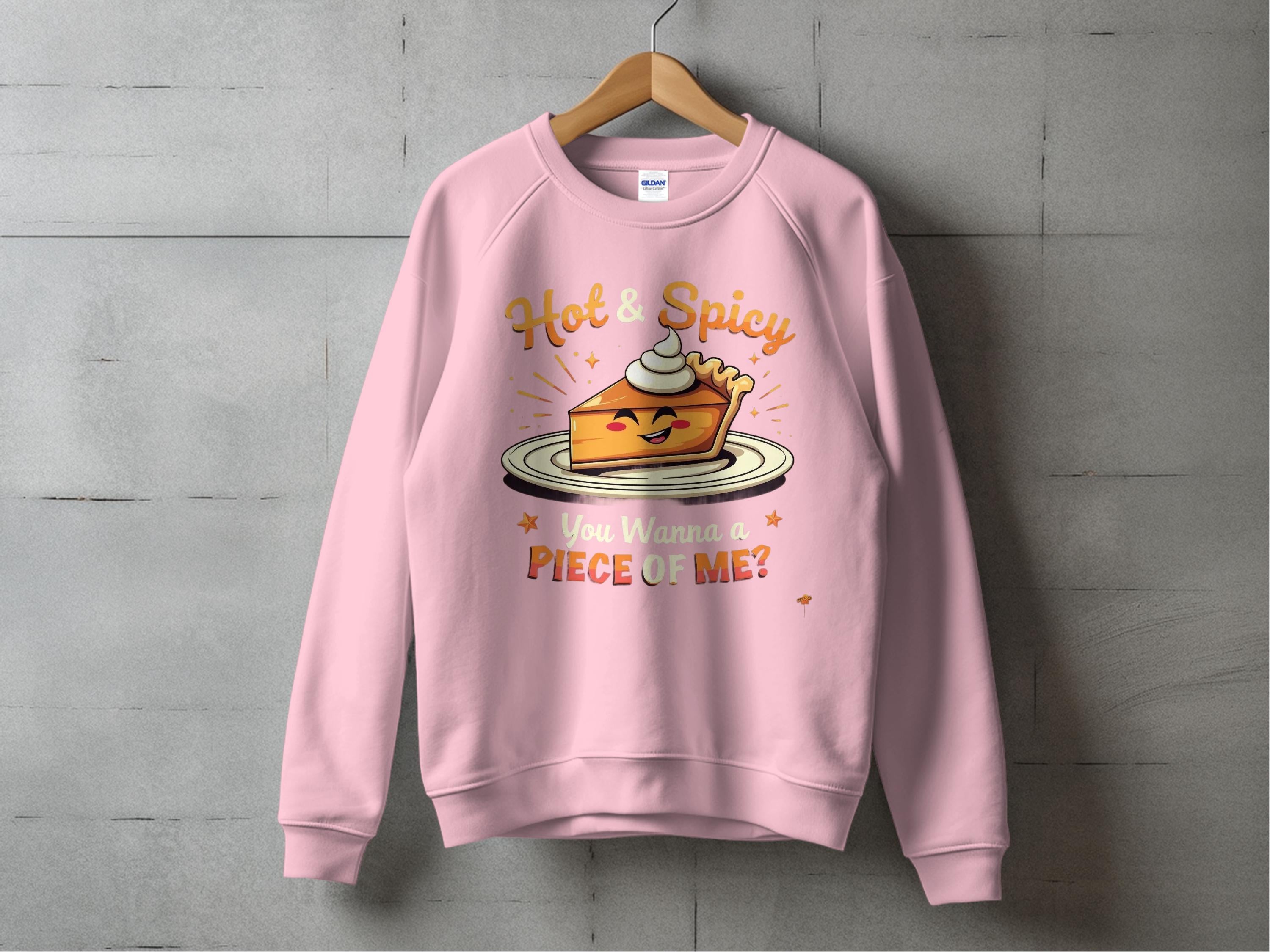 A Garment Graphics pink sweatshirt hangs against a concrete wall, showcasing a playful pie character with whipped cream, arms, and a smile. The text reads Hot & Spicy and You Wanna a Piece of Me? from the unisex Hot and Spicy Sweatshirts line.