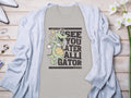 Garment Graphics presents a casual t-shirt featuring a cartoon alligator with a basketball and the phrase See You Later Alligator. Styled on wood, its paired with a light gray cardigan, flowers, and accessories for a relaxed vibe.