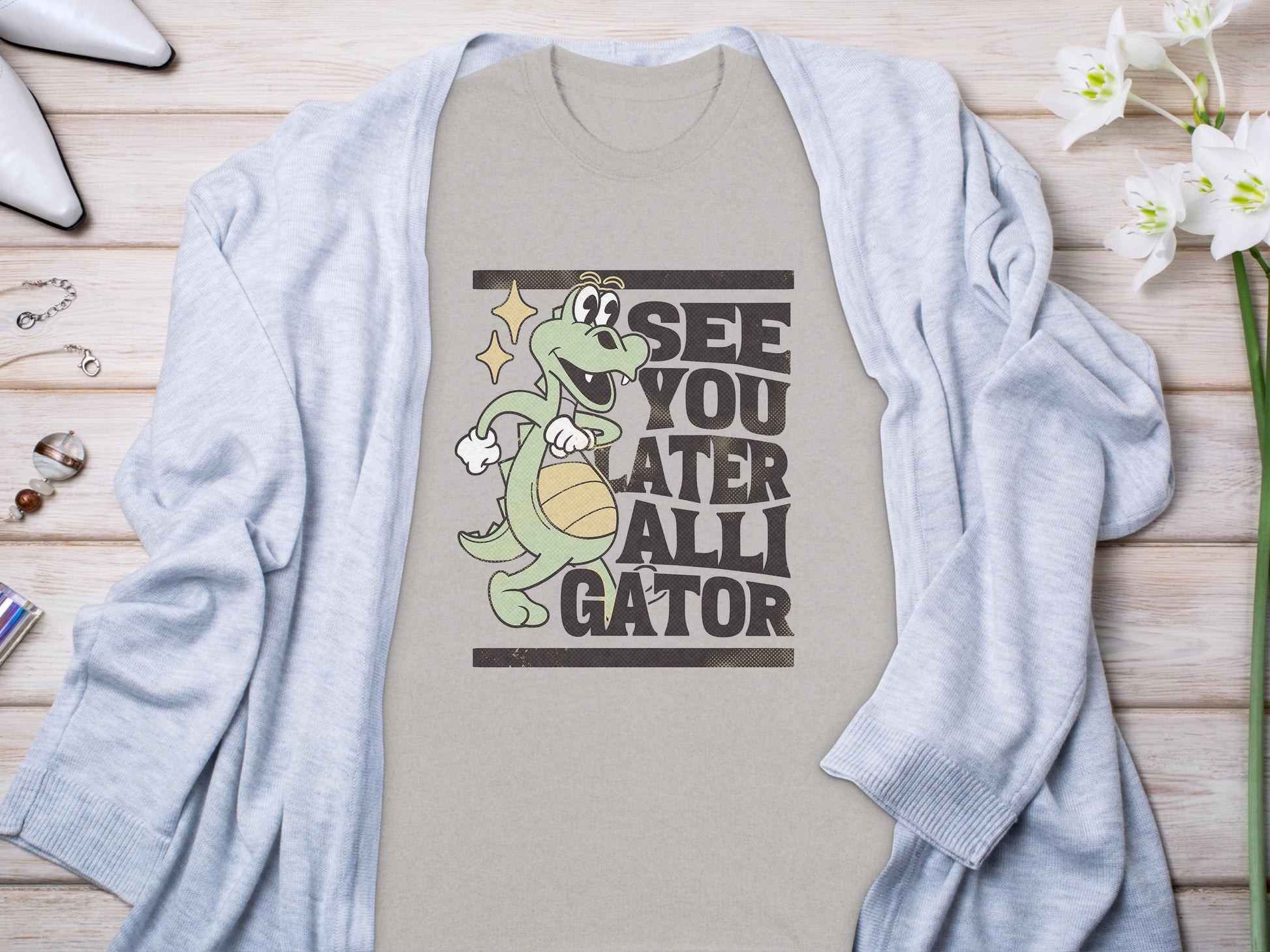 Garment Graphics presents a casual t-shirt featuring a cartoon alligator with a basketball and the phrase See You Later Alligator. Styled on wood, its paired with a light gray cardigan, flowers, and accessories for a relaxed vibe.