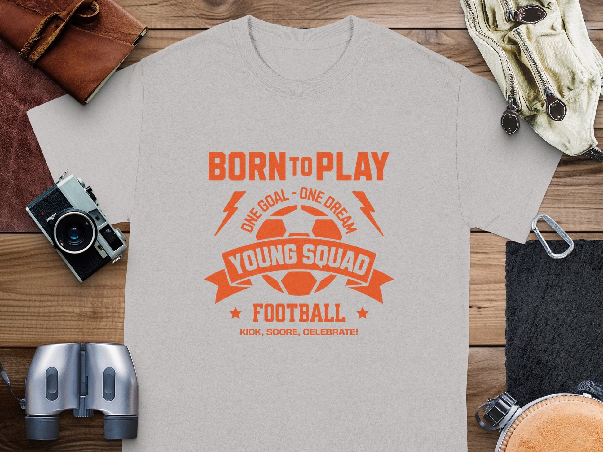 A Garment Graphics gray t-shirt with orange Born to Play, One Goal - One Dream, and Kick, Score, Celebrate graphics lays on a wooden surface. Its surrounded by a camera, binoculars, and bags, ready for the next big game.