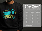 A person wears a black sweatshirt from Garment Graphics featuring the bold, colorful Take It Easy design with arrows and musical notes. Nearby, a Gildan 18000 size chart shows unisex sizes from S to 5XL with measurements in inches.
