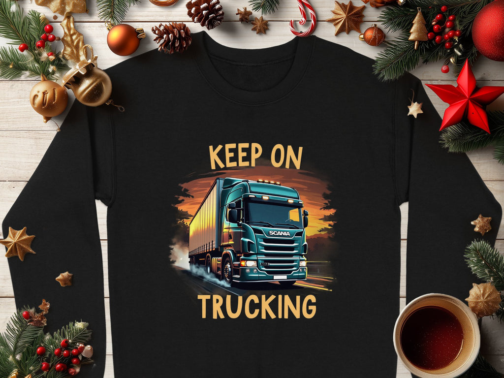 Garment Graphics black graphic sweatshirt for truck enthusiasts displays Keep On Trucking in bold yellow letters above a large blue truck illustration, surrounded by festive decorations like pine cones, baubles, and a cozy coffee cup—ideal for road life lovers.