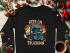 Garment Graphics black graphic sweatshirt for truck enthusiasts displays Keep On Trucking in bold yellow letters above a large blue truck illustration, surrounded by festive decorations like pine cones, baubles, and a cozy coffee cup—ideal for road life lovers.