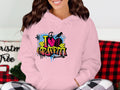 A person is wearing a light pink Garment Graphics hoodie adorned with a vibrant urban street art design stating I Love Graffiti, set against a plaid background, with hints of Christmas decoration adding whimsy to this casual classic.