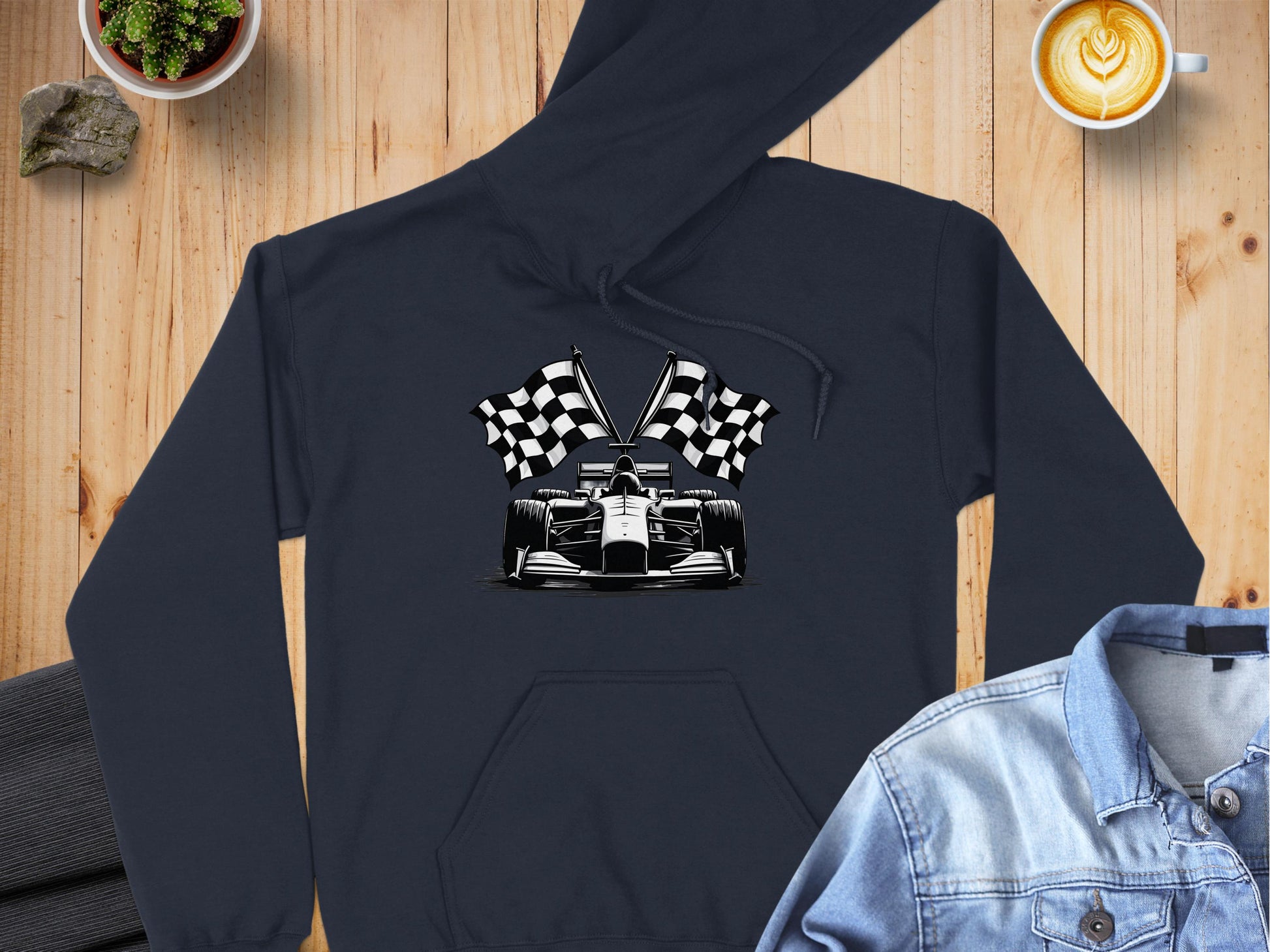 Garment Graphics presents a black racing-themed hoodie featuring a race car graphic with checkered flags. It rests on wood, complemented by a latte cup, succulent plant, folded pants, and light denim jacket.