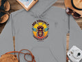 Garment Graphics gray Bee Yourself hoodie from the Summer Edition showcases a cartoon bee in sunglasses with the text Bee Yourself, Everyone Else is Taken. Pair it with a straw hat, sandals, map, notebook, and camera for an eye-catching ensemble.