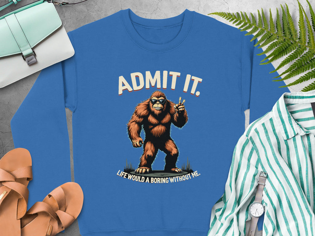 The Garment Graphics blue sweatshirt showcases a cartoon Bigfoot flashing a peace sign, with ADMIT IT. Life would be boring without me. Ideal for casual wear, it pairs well with sandals and stripes. Displayed on ferns and wood, it comes in unisex sizes.