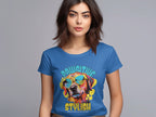 A woman with shoulder-length brown hair wears a Garment Graphics womens blue t-shirt featuring a graphic of a dog in sunglasses and the words Pawsitive Stylish on a plain gray background.