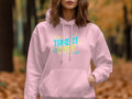 Wearing a Garment Graphics light pink hoodie emblazoned with Take It Easy in colorful text, the person creates a cozy fashion vibe, standing amidst an autumn forest backdrop with blurred trees and leaves. One hand rests casually in the hoodie pocket.