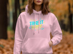 Wearing a Garment Graphics light pink hoodie emblazoned with Take It Easy in colorful text, the person creates a cozy fashion vibe, standing amidst an autumn forest backdrop with blurred trees and leaves. One hand rests casually in the hoodie pocket.