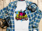 A Garment Graphics white t-shirt featuring a vibrant I Love Graffiti design with spray cans and brick patterns lies beneath a blue and green flannel shirt. Nearby, black headphones, a camera, and a white cup are neatly arranged on a wooden surface.