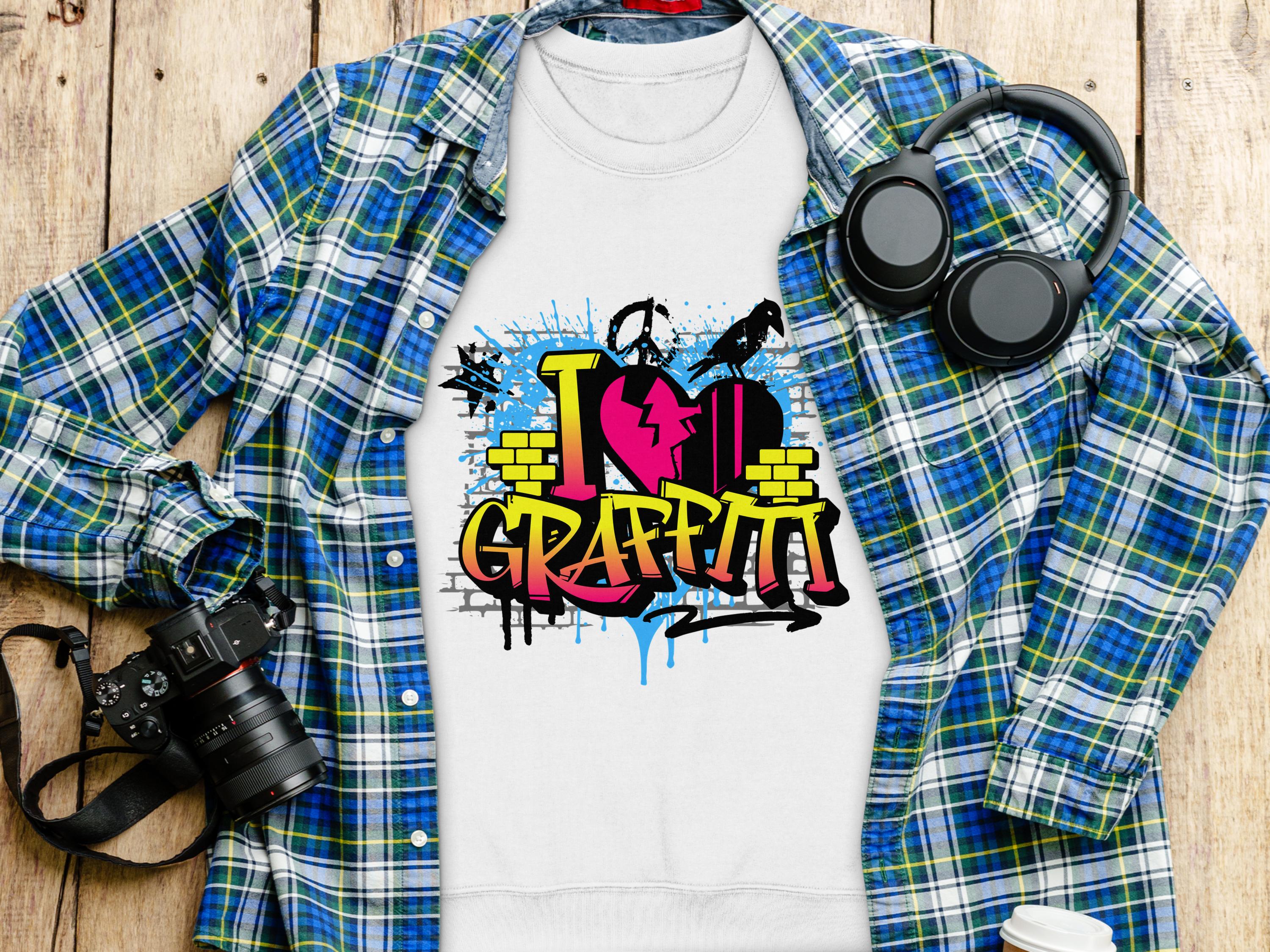 A Garment Graphics white t-shirt featuring a vibrant I Love Graffiti design with spray cans and brick patterns lies beneath a blue and green flannel shirt. Nearby, black headphones, a camera, and a white cup are neatly arranged on a wooden surface.