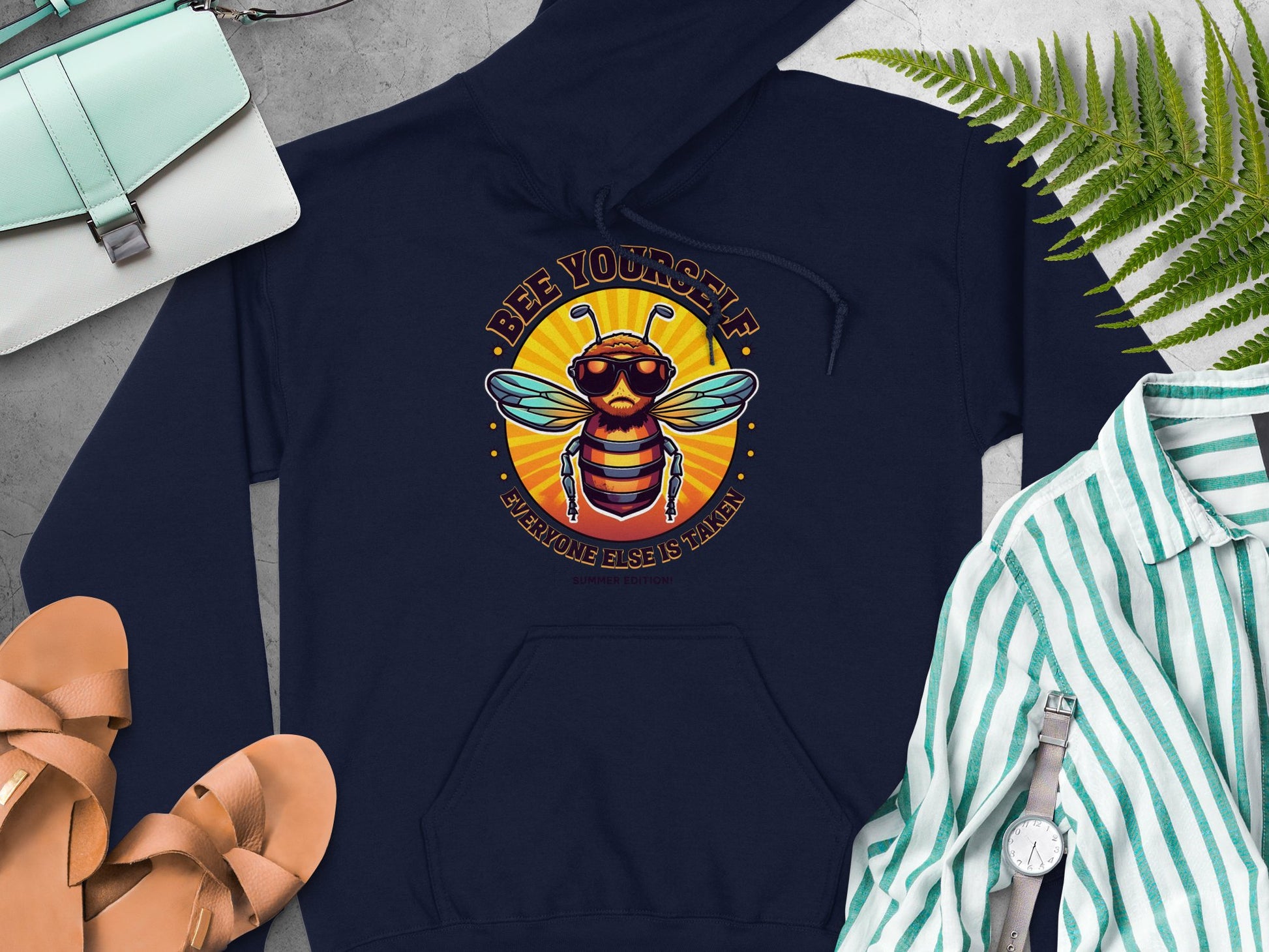 A Garment Graphics navy Bee Yourself Hoodie shows a bee in sunglasses with “Everyone Else is Taken.” Its paired with a striped shirt, tan sandals, and light gray purse on a gray surface, surrounded by green leaves for the Summer Edition.